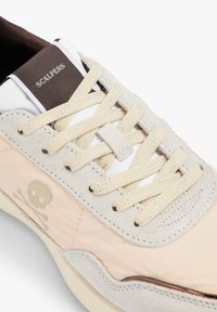 Zapatilla beige y gris claro con cordones crema, etiqueta "SCALPERS" en la lengüeta y un logo de calavera y tibias cruzadas en el panel lateral.