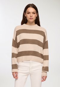 Sweter z dzianiny w paski w kolorze kremowym i brązowym, z wysokim kołnierzem i opadłymi ramionami, zestawiony z białymi dżinsami. Teksturowany materiał z prążkowanymi mankietami.