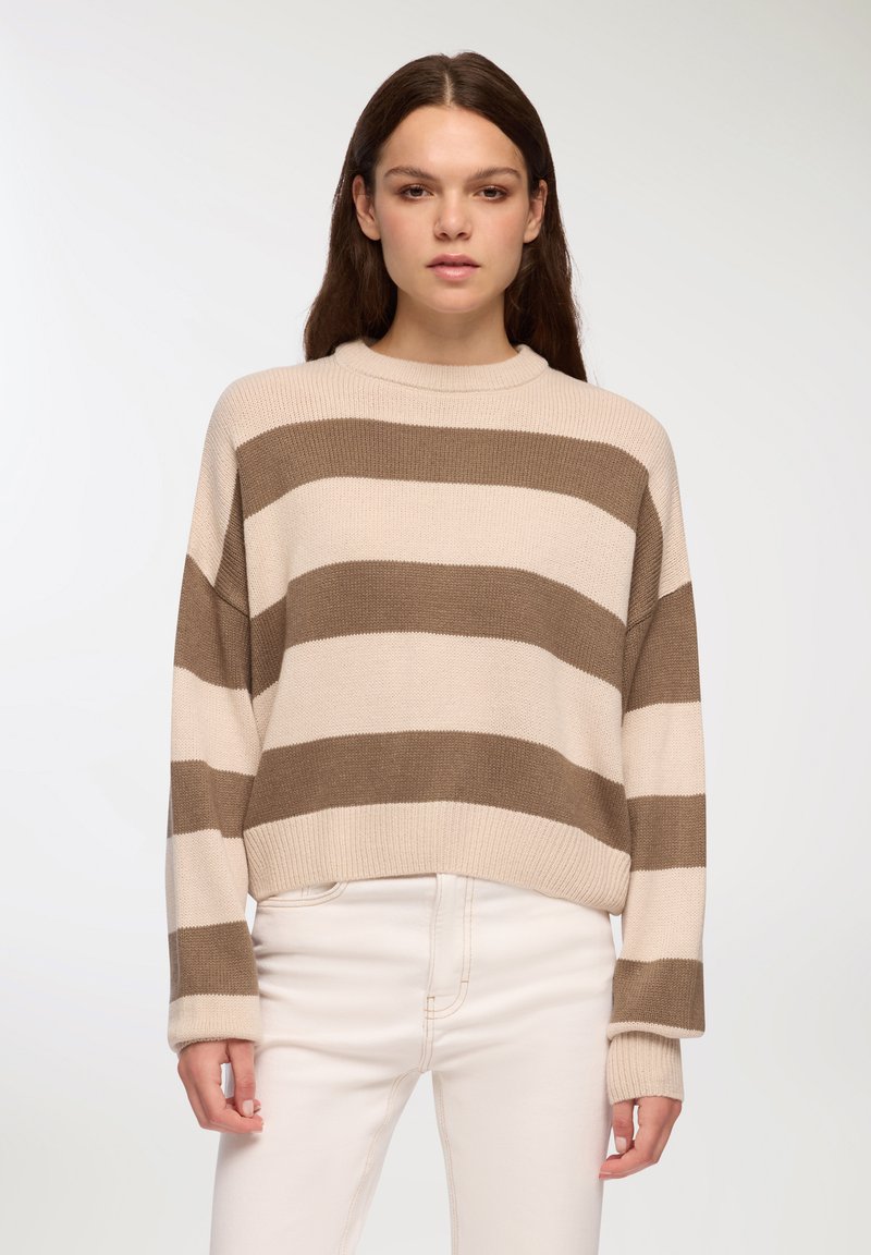 Sweter z dzianiny w paski w kolorze kremowym i brązowym, z wysokim kołnierzem i opadłymi ramionami, zestawiony z białymi dżinsami. Teksturowany materiał z prążkowanymi mankietami.