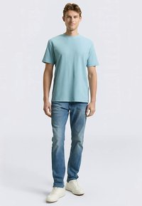 Hellblaues Baumwoll-T-Shirt mit kurzen Ärmeln, kombiniert mit mittelblauen Jeans und weißen Turnschuhen. Das Model steht aufrecht und schaut nach vorne.