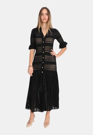 Giovane donna posa con un vestito lungo nero con bottoni, motivi in pizzo e maniche a sbuffo a metà braccio, indossa tacchi beige a punta.