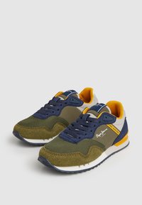 Zapatillas con una mezcla de malla verde oliva y ante azul marino, con un acento amarillo en el talón y la lengüeta, suela blanca y suela exterior de goma negra.