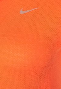 Top deportivo de Nike en un vibrante color naranja, con un tejido suave y texturizado que presenta pequeñas perforaciones y un logo plateado en el pecho.
