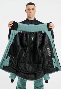 Teal waterdichte jas met zwarte binnenkant, voorzien van mesh zakken, logo-tags en een gladde buitenkant, opengehouden om de interne details te tonen.