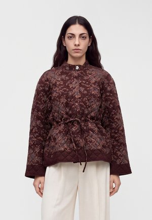 Femme aux longs cheveux foncés porte une veste matelassée bordeaux à motif floral avec une ceinture à la taille et un pantalon blanc à jambes larges, debout devant un fond uni.