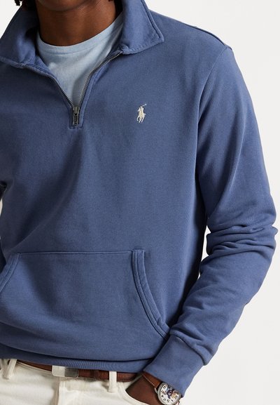 Suéter azul de peso medio con media cremallera, que presenta un cuello acanalado, bolsillo frontal y un pequeño logo bordado en el pecho. Tejido texturizado.