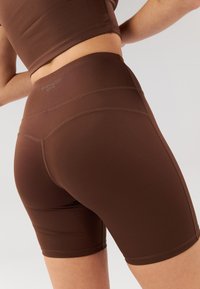 Brown high-waisted active shorts made of smooth fabric, featuring a fitted design and subtle stitching details. Brand logo is visible on the waistband.
