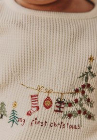 Kreminės spalvos megztinis su tekstūruotais raibėjimais, puošiamas sudėtinga kalėdinių tematikos siuvinėjimu, įskaitant kojines, papuošimus ir medelius.