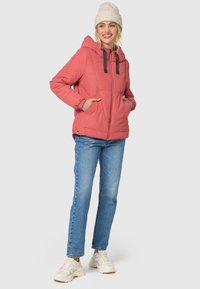 Koralfarvet pufferjakke med hætte, elastiske manchetter og to forlommer. Sammen med lyseblå jeans og hvide sneakers.