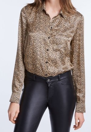 Kobieta nosząca bluzkę z długim rękawem w panterkę, wpuszczoną do czarnych skórzanych spodni z wysokim stanem, stojąca na jednolitym tle.