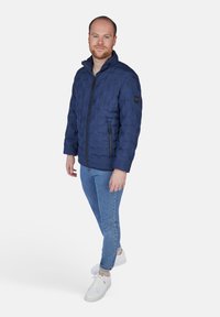 Giacca imbottita blu navy con design trapuntato, chiusura frontale con zip e tasche laterali. Indossata sopra una camicia leggera e abbinata a jeans blu e sneakers bianche.