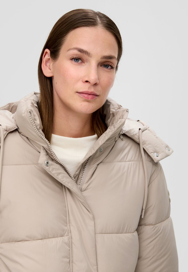 Hellebeige gepolsterte Jacke mit hohem Kragen, Reißverschluss vorne und Kordelzugkapuze. Merkmale sind metallische Hardware und eine gesteppten Textur.