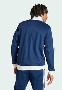Marineblaue Sportjacke mit weißen Akzenten und elastischem Bund, mit hohem Kragen und langen Ärmeln, von hinten gezeigt.