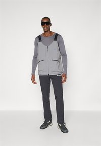 Gray lightweight vest with zip closure, shoulder straps, and multiple front pockets. Paired with black pants and gray athletic shoes.