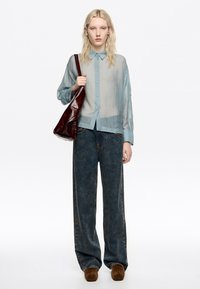 Przezroczysta, jasnoniebieska koszula z guzikami i kołnierzykiem, w połączeniu z luźnymi, ciemnoniebieskimi dżinsami i brązowymi zamszowymi butami. Trzyma bordową skórzaną torbę.