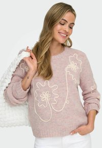 Kvinne som smiler, iført en rosa strikkegenser med kremfarget blomstermønster, hvite bukser, holder en teksturert hvit veske over skulderen.