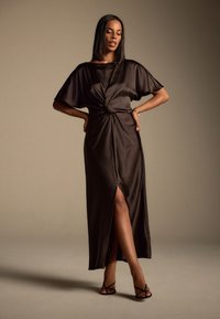 Brun satin maxi-kjole med korte ærmer, knudetaljedetalje og lårhøjt slids. Stylet med sorte stropper og høje hæle. Glat tekstur.