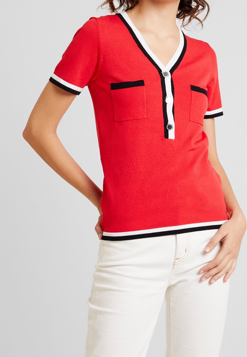 Femme portant un haut rouge à manches courtes avec un bord noir et blanc, deux poches avant, des boutons blancs et un pantalon blanc.