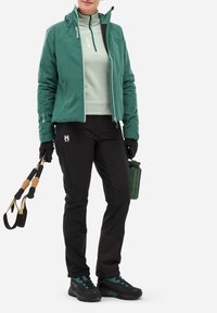 Veste imperméable verte avec une fermeture éclair à l'avant, associée à un pantalon noir et des bottes de randonnée noires avec des accents turquoise. Tient des bâtons de trekking et une bouteille d'eau verte.