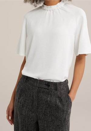 Vrouw die een wit blouse met korte mouwen en gerimpelde kraag draagt, ingestopt in een grijze, hooggetailleerde broek met textuur, hand in de zak.