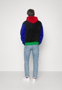 Měkká mikina s barevnými bloky: černý hlavní díl, červená kapuce, modré rukávy, zelený lem. Nosená s světle modrými džínami a bílými teniskami, viditelné vyhrnuté manžety.