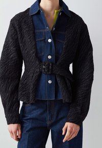 Svart texturerad jacka med bältesliv och öppen framsida, lager på lager över en denimskjorta med ljusblå knappar och gröna accenter.
