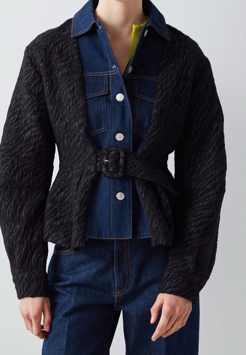 Svart texturerad jacka med bältesliv och öppen framsida, lager på lager över en denimskjorta med ljusblå knappar och gröna accenter.