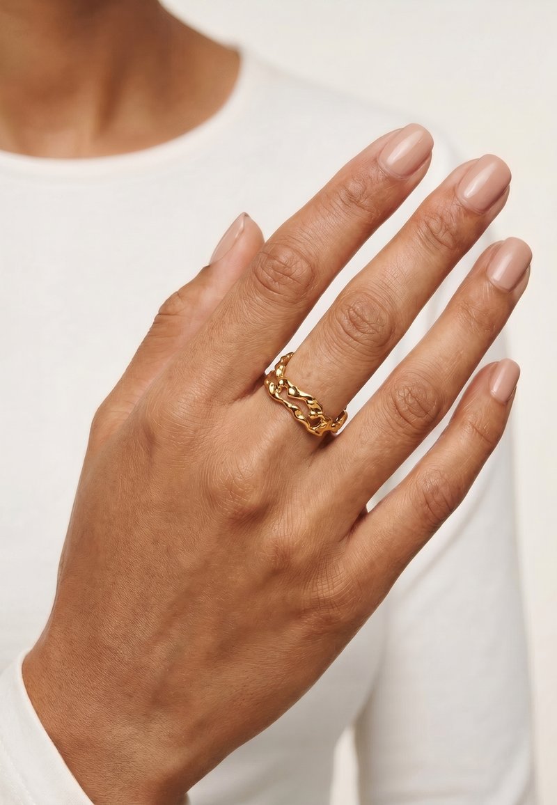 Main avec peau brun moyen portant une bague dorée texturée au majeur sur fond de vêtements blancs.