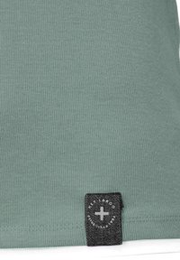 T-shirt in cotone a coste di colore verde chiaro, con un'etichetta in tessuto nera che presenta un logo circolare e del testo; texture liscia e design aderente.