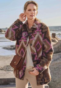 Veste en laine à motifs marron et rose avec col et boutons, accompagnée d'un haut clair et d'un pantalon beige, avec un sac bandoulière marron.