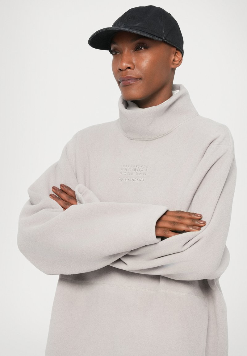 Persoon in een lichtgrijze hoge hals sweatshirt met een logo, een zwarte voetbalpet, met gekruiste armen, die iets opzij kijkt tegen een effen achtergrond.