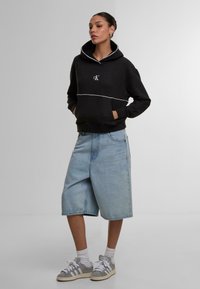 Schwarzer Kapuzenpullover mit weißen Akzentnähten, Fronttasche, kombiniert mit hellblauen Jeansshorts und grauen Turnschuhen. Das Model steht vor einem schlichten Hintergrund.