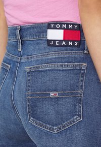 Jeans in denim blu scuro con un'etichetta con logo e dettagli in rosso, bianco e blu navy sulla vita, e un piccolo logo sulla tasca posteriore.