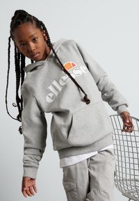 Sudadera gris con un bolsillo delantero, que presenta un logo blanco y un acento colorido. El modelo lleva pantalones de color claro y posa junto a una silla de alambre.