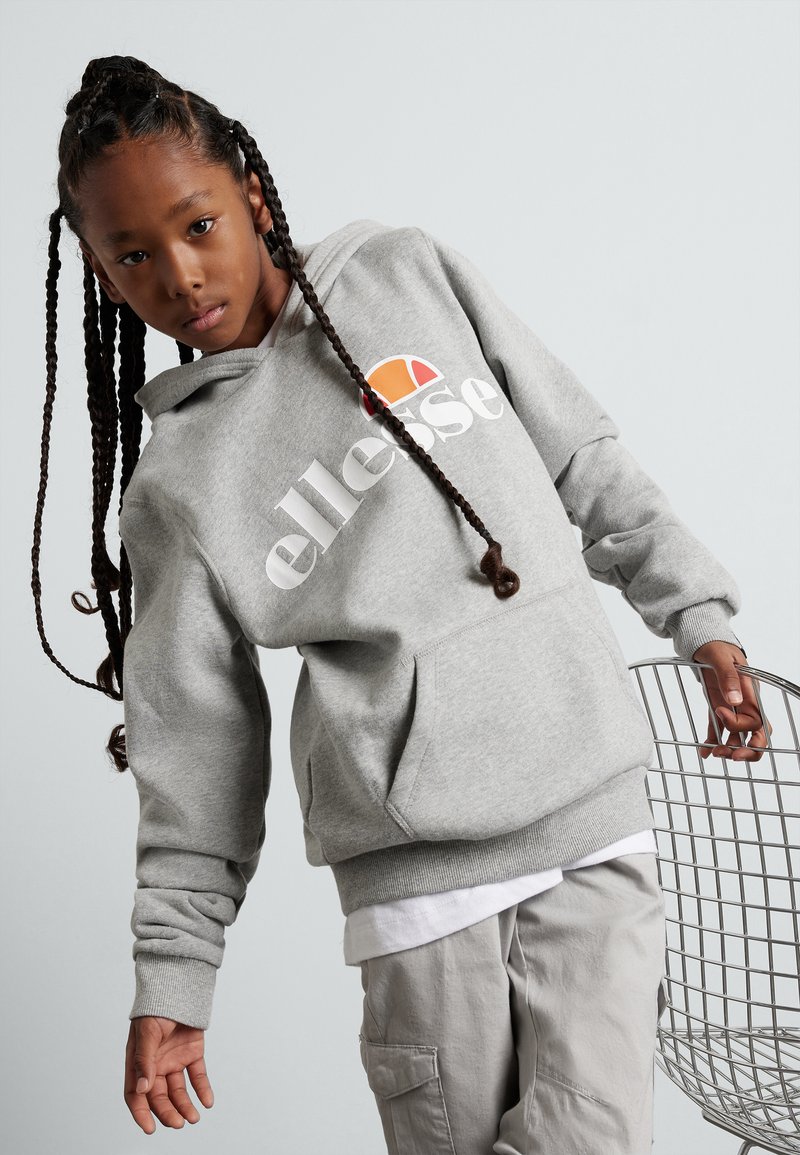 Sudadera gris con un bolsillo delantero, que presenta un logo blanco y un acento colorido. El modelo lleva pantalones de color claro y posa junto a una silla de alambre.