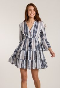 Navy und weiß gemustertes Kleid mit ausgestellten Ärmeln, V-Ausschnitt und Schlaufen-Detail in der Taille. Verfügt über einen gestuften Rüschensaum und Quastenakzente.