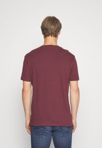 Bordeauxrotes T-Shirt mit kurzen Ärmeln und rundem Ausschnitt; aus weichem Stoff gefertigt. Kombiniert mit blauen Jeans, was ein schlichtes, lässiges Design zeigt.