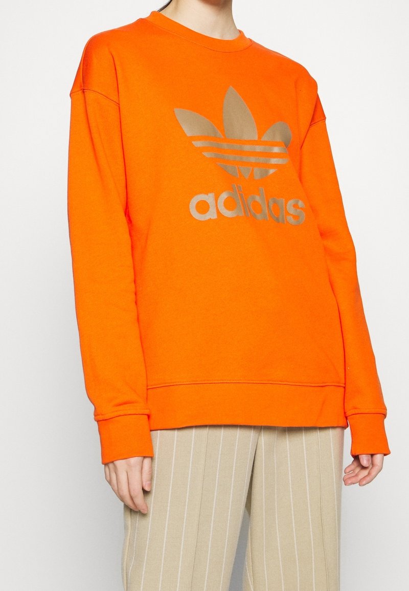 Orange sweatshirt med guld Adidas-logotyp och grafik med tre ränder. Har långa ärmar och ribbad mudd, tillverkad av mjukt material.