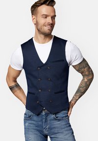 Granatowa kamizelka dwurzędowa z brązowymi guzikami i dwiema przednimi kieszeniami, noszona na białej koszulce, zestawiona z niebieskimi dżinsami.