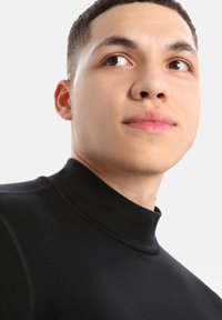Fekete, magas nyakú felső, sima textúrával és testhez simuló fazonnal, kiemelve a tiszta vonalakat és a modern stílust. Rövid haj és semleges háttér.