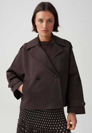 Frau mit kurzen dunklen Haaren, die eine dunkelbraune, zweireihige Jacke und einen schwarzen Rock mit weißen Punkten trägt, steht vor einem einfarbigen Hintergrund.