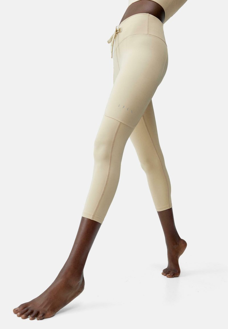 Bež leggins s prilegajočo zasnovo, dolžine do sredine tele, in pasom z vrvico. Imajo brezšivne šive in diskretno logotip na strani.
