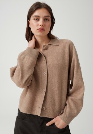 Giovane donna con capelli castani indossa un cardigan beige con bottoni e pantaloni neri, una mano tocca il collo, l’altra nella tasca, su sfondo chiaro.