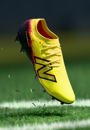 Gul og sort fodboldstøvle med røde detaljer og bogstavet "N" i luften over en græsmark med en hvid linje.