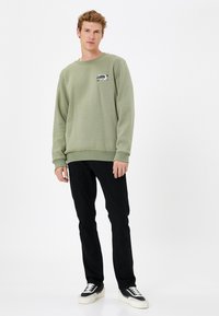 Sudadera verde claro con cuello redondo, puños de canalé y detalle de logo bordado, combinada con pantalones negros y zapatillas negras.