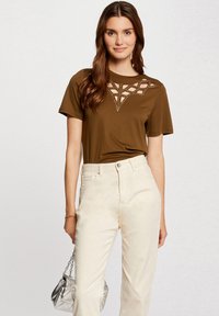 Brązowa koszula z krótkim rękawem o wyciętym geometrycznym wzorze i ćwiekami przy dekolcie, w połączeniu z jasnymi spodniami i srebrną torbą.