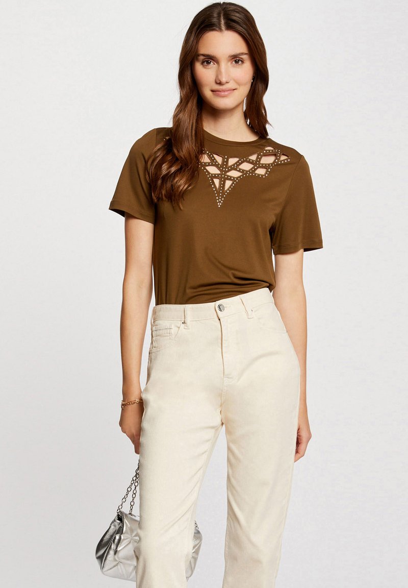 Brązowa koszula z krótkim rękawem o wyciętym geometrycznym wzorze i ćwiekami przy dekolcie, w połączeniu z jasnymi spodniami i srebrną torbą.