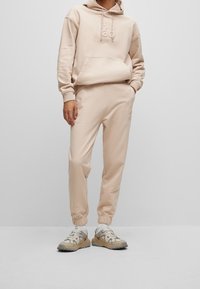 Felpa beige con grande tasca frontale e logo, abbinata a joggers tapered coordinati. Le scarpe presentano una combinazione di tonalità beige e grigie.