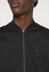 Gros plan d'une personne portant une veste noire zippée avec un col côtelé et le logo "BOSS" sur la poitrine.