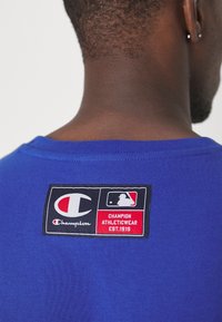 Modrá bavlněná tričko s černou a červenou nášivkou obsahující loga Champion a MLB, umístěné na zadní straně blízko výstřihu.